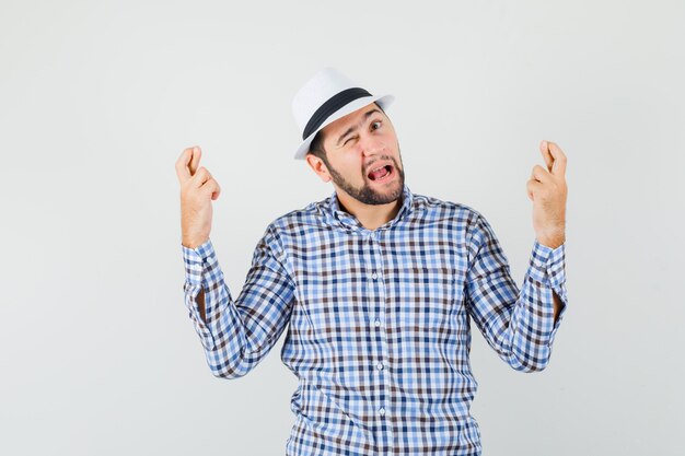 Young male in checked shirt, hat keeping fingers crossed, opening mouth, winking eye , front view.