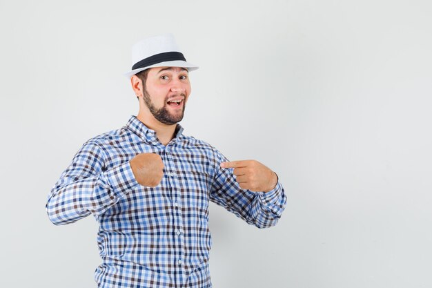 Young male in checked shirt, hat pointing fingers at himself and looking proud , front view.