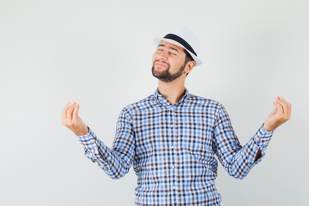 Young male in checked shirt, hat showing italian gesture and looking peaceful , front view.