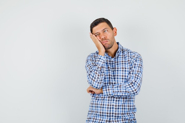 Young male in checked shirt leaning cheek on raised palm and looking hesitant