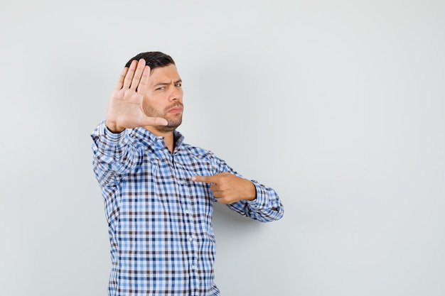 Young male in checked shirt showing stop gesture by pointing at himself and looking confident