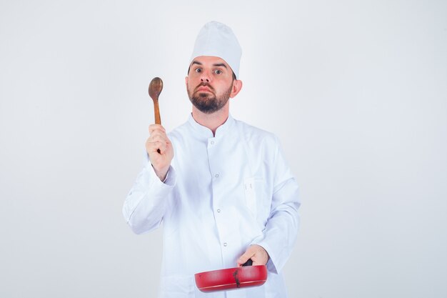 Young male chef holding frying pan and wooden spoon in white uniform and looking thoughtful. front view.