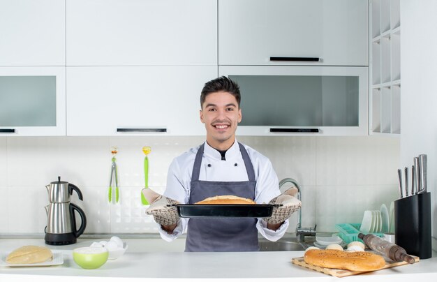 Young male chef in uniform standing behind table wearing holder and holding freshly-baked bread in the white kitchen