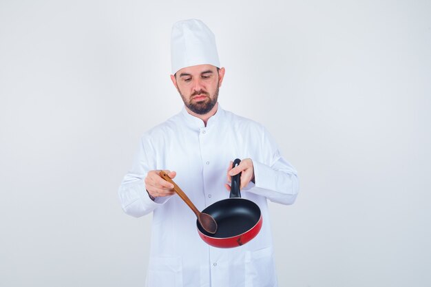 Молодой шеф-повар-мужчина в белой форме держит пустую сковороду деревянной ложкой и смотрит вниз, вид спереди.