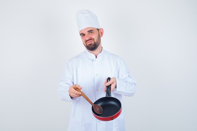 Young male chef in white uniform showing empty frying pan with wooden spoon and looking disappointed , front view.