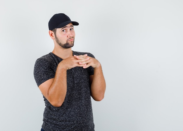 Young male clasping fingers on chest in black t-shirt and cap and looking indecisive