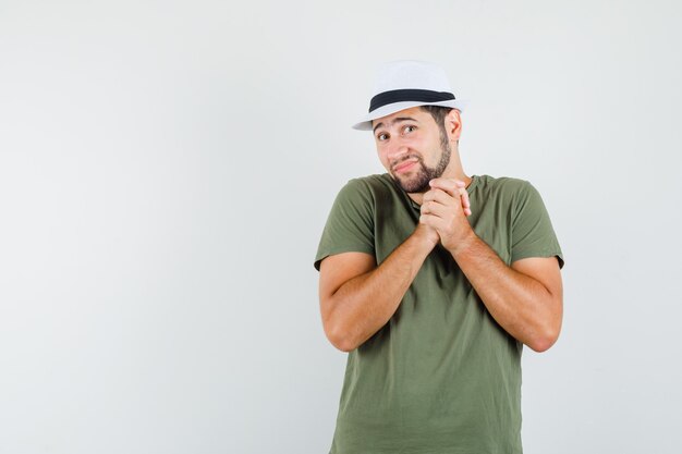 Young male clasping hands in praying gesture in green t-shirt and hat and looking hopeful