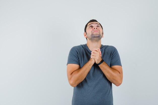 Young male clasping hands in praying gesture in grey t-shirt and looking hopeful 