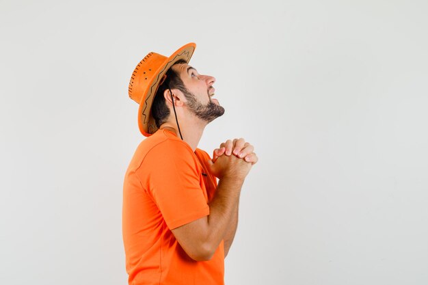 Young male clasping hands in praying gesture in orange t-shirt, hat and looking hopeful .