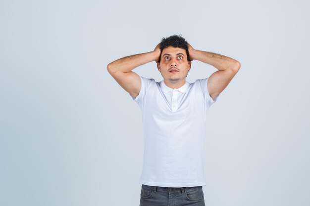 Young male clasping head in hands in white t-shirt, pants and looking helpless , front view.