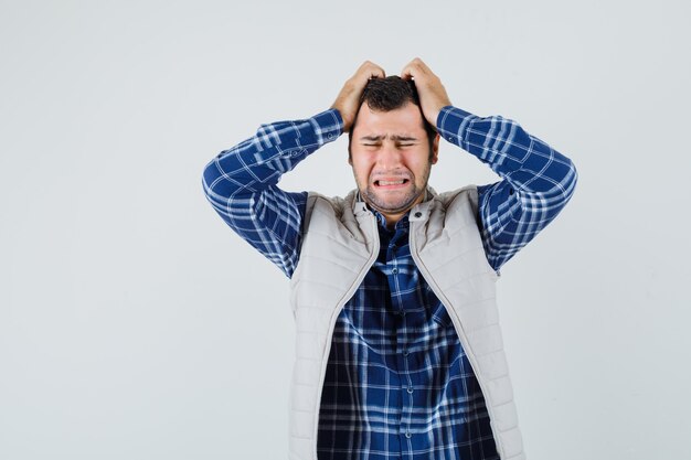 Young male clasping head with hands in shirt,sleeveless jacket and looking stressful. front view.