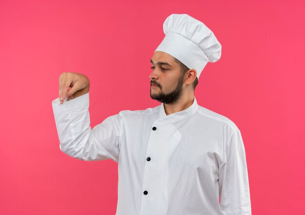 Young male cook in chef uniform adding salt and looking at side isolated on pink space