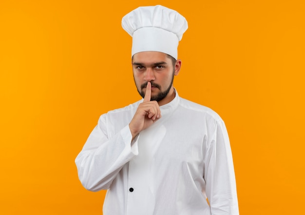 Young male cook in chef uniform gesturing silence isolated on orange space