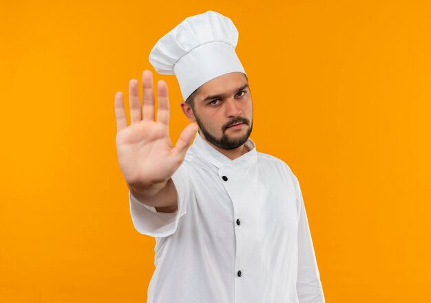 Young male cook in chef uniform gesturing stop  isolated on orange space