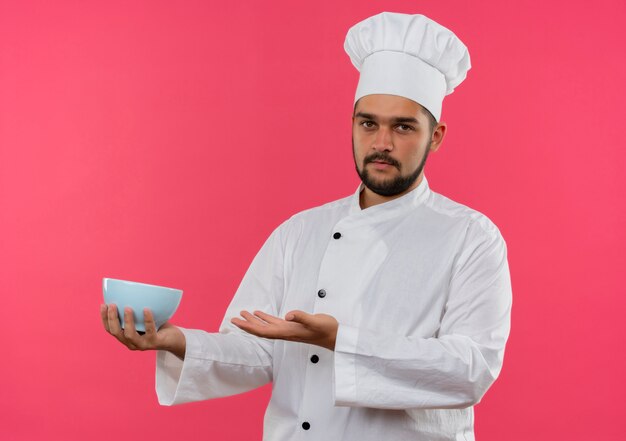 Young male cook in chef uniform holding bowl isolated on pink space