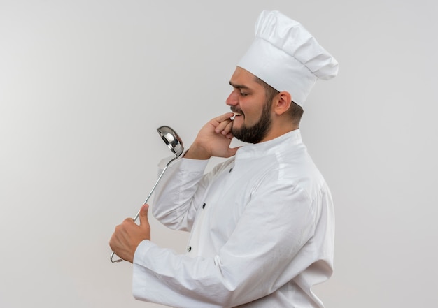 Young male cook in chef uniform holding ladle and using it as mirror isolated on pink space 