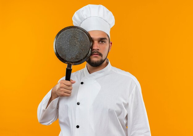 Young male cook in chef uniform holding and looking from behind frying pan isolated on orange space