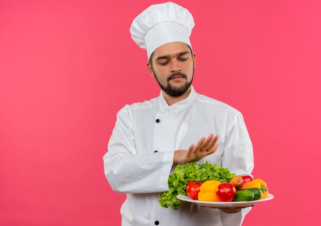 Young male cook in chef uniform holding and looking at plate of vegetables and keeping hand above plate isolated on pink space 