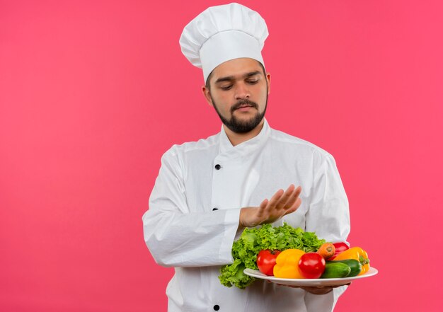 Молодой мужчина-повар в униформе шеф-повара держит и смотрит на тарелку с овощами и держит руку над тарелкой, изолированной на розовом пространстве