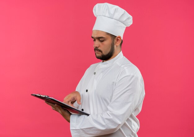 Young male cook in chef uniform holding looking at and putting finger on clipboard isolated on pink space 