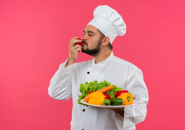 Giovane cuoco maschio in uniforme del cuoco unico che tiene piatto di verdure e annusando il pomodoro con gli occhi chiusi isolati su spazio rosa