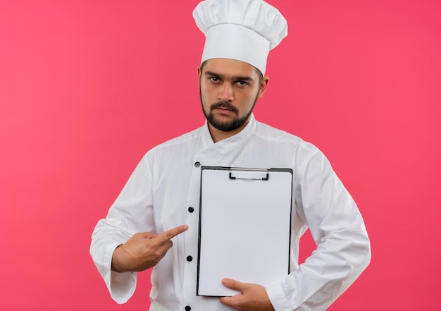 요리사 유니폼 잡고 클립 보드를 가리키고 분홍색 공간에 고립 된 찾고 젊은 남성 요리사