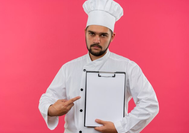 Young male cook in chef uniform holding and pointing at clipboard and looking  isolated on pink space