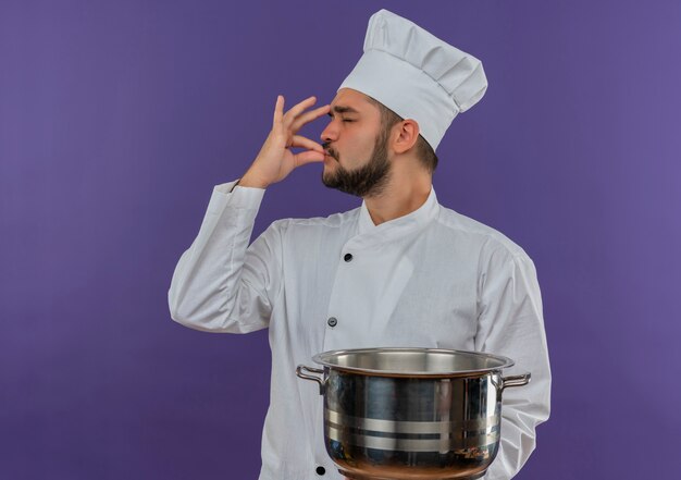 Giovane cuoco maschio in uniforme del cuoco unico che tiene pentola e facendo gesto gustoso isolato su spazio viola