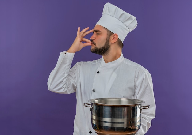 Молодой мужчина-повар в униформе шеф-повара держит горшок и делает вкусный жест, изолированный на фиолетовом пространстве
