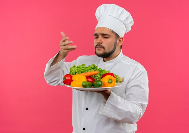 Young male cook in chef uniform holding and sniffing plate of vegetables with closed eyes isolated on pink space