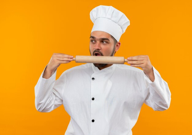 요리사 유니폼 잡고 롤링 핀을 물고 오렌지 공간에 고립 된 측면을보고 젊은 남성 요리사