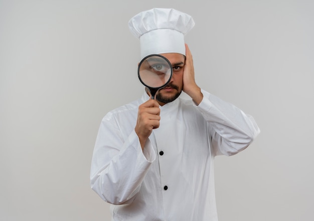 Молодой мужчина-повар в униформе шеф-повара смотрит через увеличительное стекло и кладет руку на голову