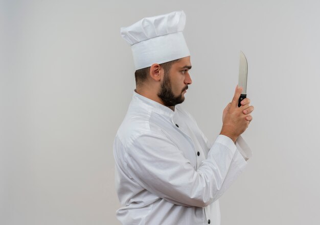 ナイフを持って見ている縦断ビューで立っているシェフの制服を着た若い男性料理人