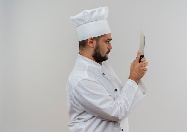 Young male cook in chef uniform standing in profile view holding and looking at knife 