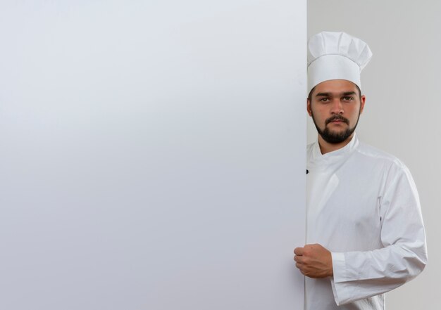 握りこぶしで見ている白い壁の後ろに立っているシェフの制服を着た若い男性料理人