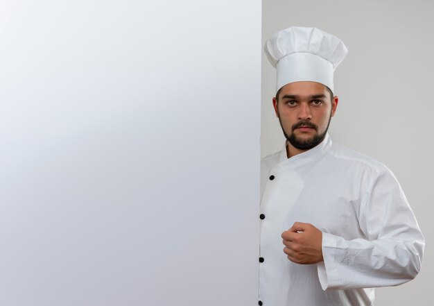 Young male cook in chef uniform standing behind white wall looking  