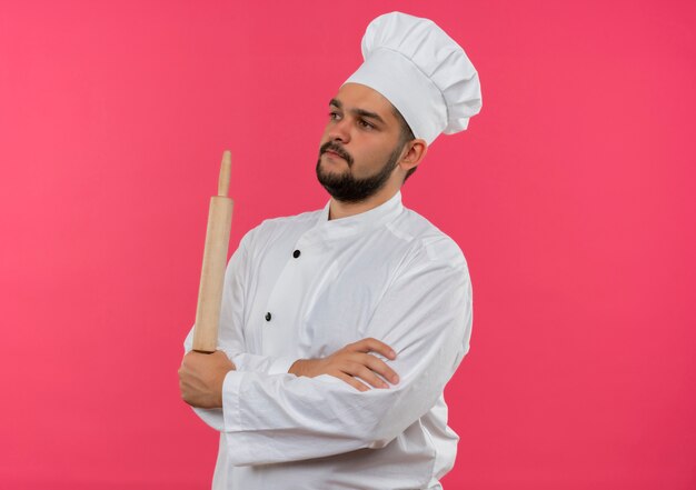 Молодой мужчина-повар в униформе шеф-повара стоит в закрытой позе и держит скалку, глядя в сторону
