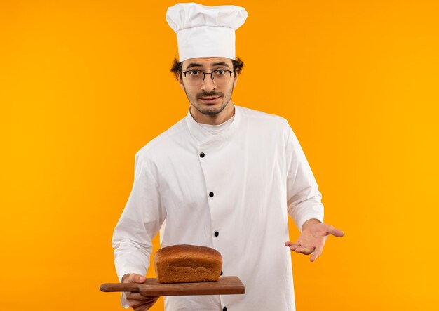 young male cook wearing chef uniform and glasses holding bread on cutting board 