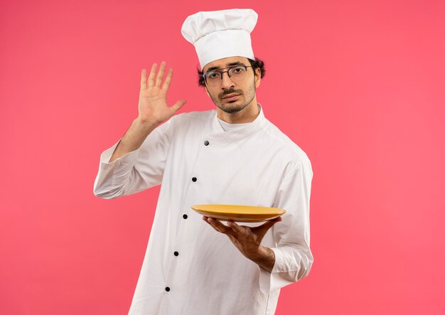 young male cook wearing chef uniform and glasses holding plate and raising hand 
