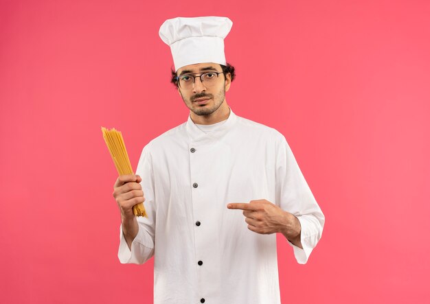 young male cook wearing chef uniform and glasses holding and points to spaghetti 