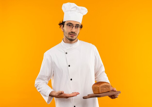 young male cook wearing chef uniform and glasses holding and points with hand to bread on cutting board 