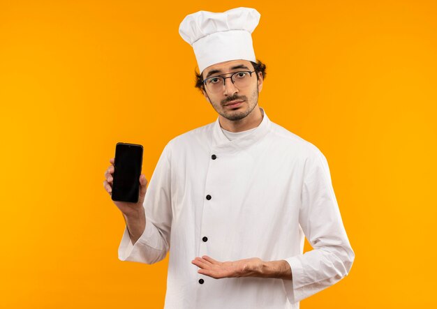 young male cook wearing chef uniform and glasses holding and points with hand to phone 