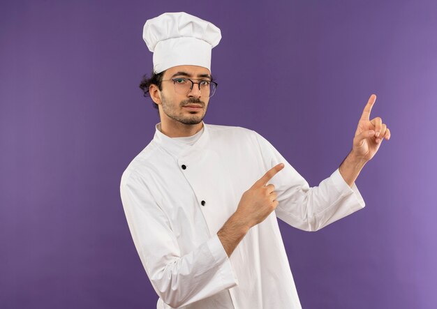 young male cook wearing chef uniform and glasses points to side 