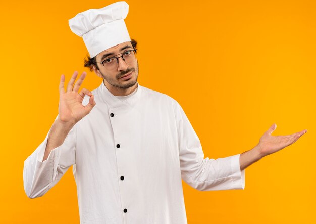 young male cook wearing chef uniform and glasses showing okey gesture and points with hand to side 