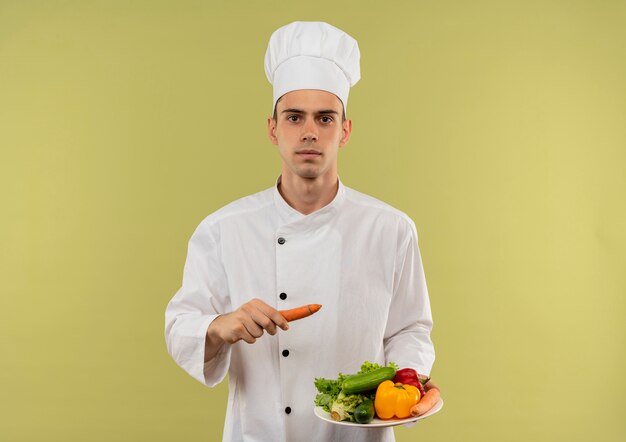 молодой мужчина-повар в униформе шеф-повара держит овощи на тарелке и морковь на изолированной зеленой стене с копией пространства