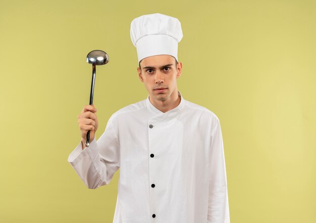  young male cook wearing chef uniform raising ladle in his hand on isolated green wall with copy space