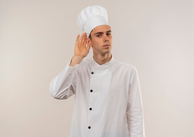  young male cook wearing chef uniform showing listen gesture on isolated white wall with copy space