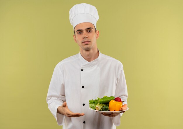  young male cook wearing chef uniform showing vegetables on plate in his hand on isolated green wall with copy space