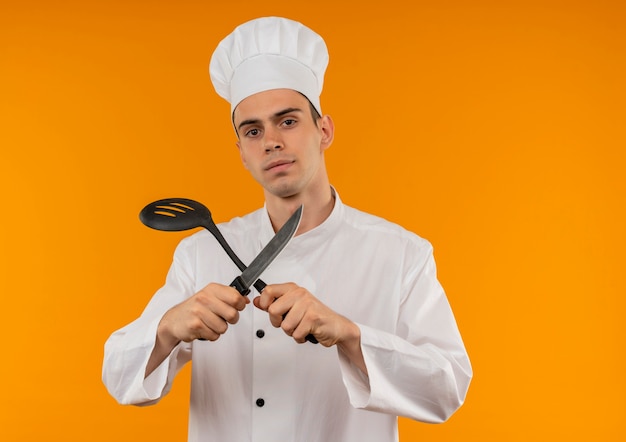  young male cool wearing chef uniform crossing ladle and knife in his hand on isolated yellow wall