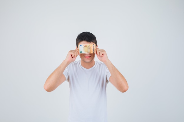 Young male covering eye with euro banknote in t-shirt and looking cheery. front view.